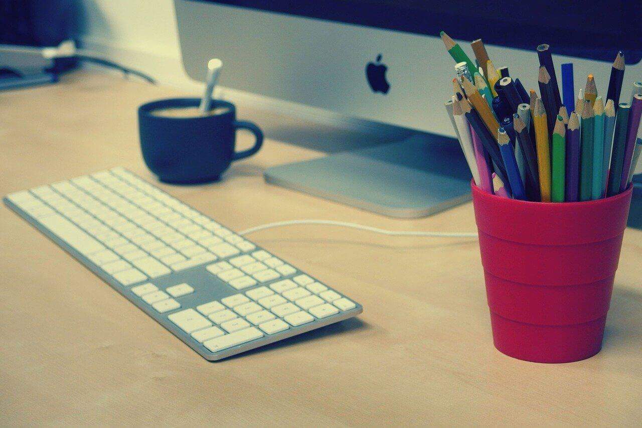 A desk with an Apple computer, cup of pencils, and a cup of coffee